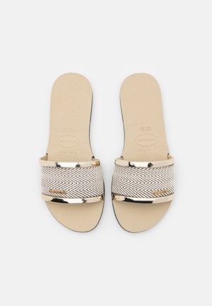 Sort-hvide mønstrede slide-sandaler med en tekstureret sål, metalliske guldfarvede detaljer og en bred rem over en beige fodseng.