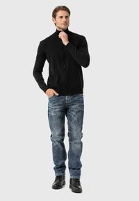 Schwarze Zip-up-Pullover mit hohem Kragen, leichtes Strickmaterial, kombiniert mit blauen Jeans mit einem ausgewaschenen Muster und weißen Nähten, schwarze Schuhe.