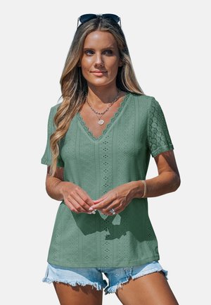 Frau mit langen blonden Haaren, die ein grünes Spitzen-Top mit kurzen Ärmeln und V-Ausschnitt trägt, geschichtete Halsketten, Sonnenbrille auf dem Kopf und helle Denim-Shorts.
