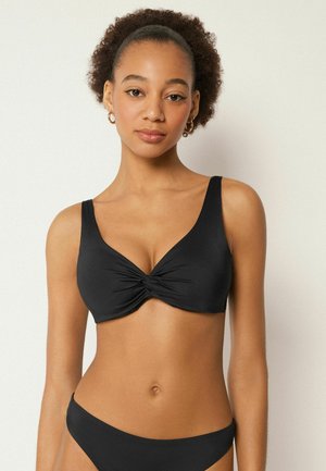 Jeune femme aux cheveux bouclés portant un bikini noir avec un détail torsadé sur le haut, debout devant un fond clair uni.