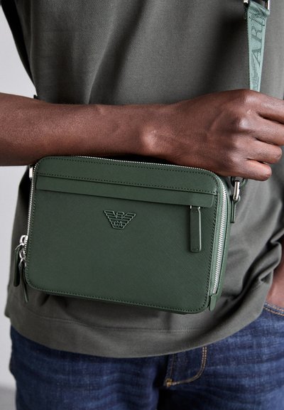 Grønn crossbody-veske laget av teksturert materiale, med glidelås, logo-akse nt og justerbar stropp. Kompakt rektangulært design.
