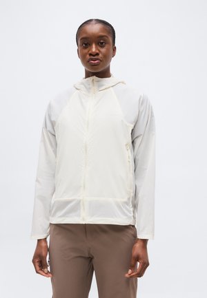 Νεαρή μαύρη γυναίκα που φοράει λευκό φούτερ με φερμουάρ και καφέ παντελόνι, στέκεται μπροστά σε απλό ανοιχτό φόντο.