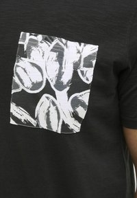 T-shirt noir avec un gros plan d'une poche rectangulaire blanche ornée d'un motif de feuille abstrait en noir et blanc, à manches courtes et texture douce.