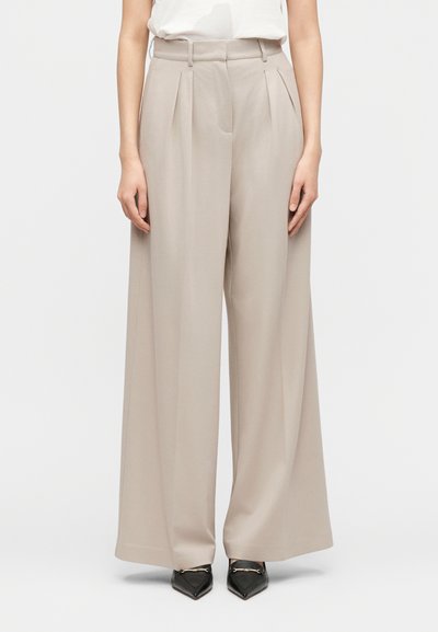 Pantalon beige taille haute à jambes larges avec plis, porté avec un haut blanc rentré et des mocassins noirs pointus avec des détails dorés.