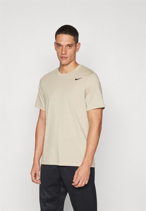 T-shirt en coton beige avec des manches courtes, un col rond et un logo Nike noir sur la poitrine. Porté avec un pantalon sombre.