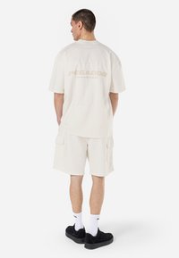 Lys beige oversized t-shirt med "PEGADOR" logo på ryggen, parret med matchende shorts med sidelommer og sorte sneakers.