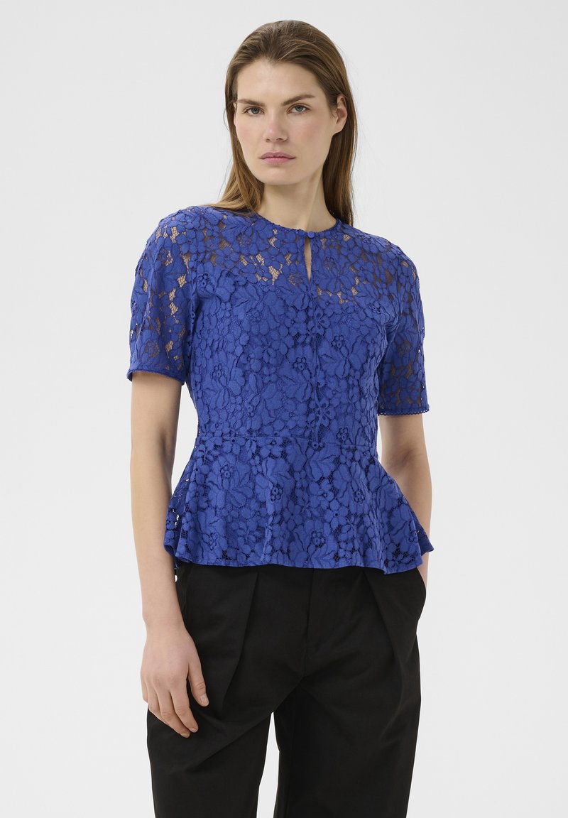 Femme portant un top ajusté en dentelle florale bleue à manches courtes et un pantalon noir, debout devant un fond clair uni.