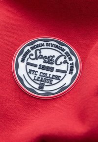 Tissu rouge avec un patch circulaire en caoutchouc noir et blanc affichant "Sport and Co." ainsi que "NYC College League 1985", avec du texte embossé et des accents.