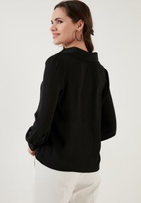 Black, long-sleeve blouse with a collar; smooth texture and tailored fit. Details include buttoned cuffs and a curved hem at the back.