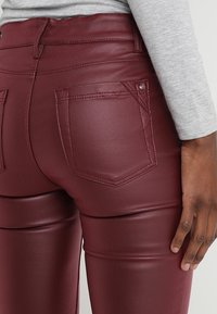Primer plano de una persona que lleva pantalones de cuero sintético burdeos con bolsillos traseros y una camiseta gris de manga larga, con la mano descansando al costado.