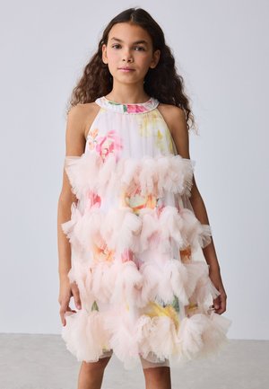 Ragazza con capelli lunghi e ricci che indossa un vestito bianco senza maniche con motivi floreali rosa e gialli e dettagli con balze stratificate.
