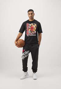 Schwarzes T-Shirt mit grafischem Design, das das Chicago Bulls-Logo zeigt, kombiniert mit schwarzen Sweatpants, die den Text "Chicago Bulls" tragen. Weiße Sneaker.
