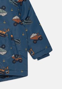 Tmavě modrá dětská bunda s rukávem, na kterém je vzor oranžových a černých stavebních vozidel a dopravních kuželů, včetně sklápěčů a rypadel.