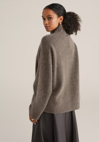 Sieviete ar curly matiem, kas sasieta augstā astē, ģērbjas taupe krāsā pūkainā turtleneck džemperī un melnās ādas svārkos, skatoties sānis pret bēšo fonu.