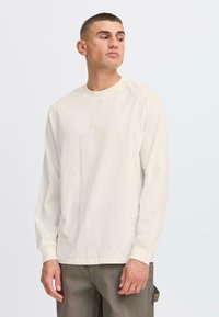 Camiseta de manga larga en color beige claro, confeccionada en algodón suave. Presenta un cuello redondo, corte relajado y puños acanalados, con un diseño minimalista.
