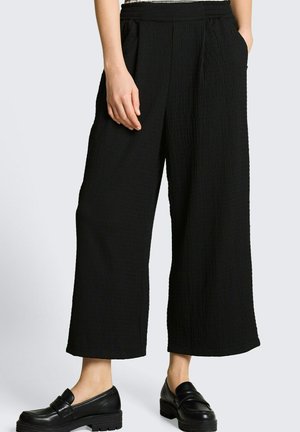Osoba nosząca czarne teksturowane szerokie spodnie o skróconej długości z elastycznym pasem i czarne, wsuwane, masywne mokasyny.