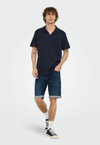 Navy blue polo shirt with a pocket, paired with rolled denim shorts, white socks, and navy blue sneakers with cream accents.