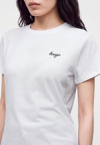 Jeune personne portant un T-shirt blanc uni à manches courtes avec un petit logo "hugo" noir en écriture cursive sur la poitrine gauche.