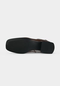 Semelle d'une chaussure habillée en cuir marron avec un talon carré et une semelle en caoutchouc noir texturé pour une bonne adhérence sur un fond clair uni.