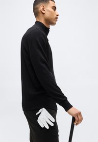 Άνδρας με μαύρο πουλόβερ και παντελόνι, κρατώντας μαύρο μπαστούνι, με λευκό γάντι στη τσέπη του, στραμμένος προς τα δεξιά, σε απλό φόντο.