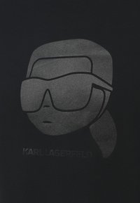 Melnā T-kreklā ir atainots pelēks stilizēts attēls ar personāžu brillēs. Zem attēla ir izdrukāts "KARL LAGERFELD" atbilstošā pelēkā krāsā.