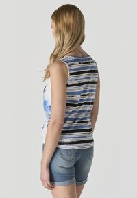 Vrouw met lang blond haar, gekleed in een mouwloos gestreept shirt en denimshorts, staand tegen een lichtgrijze achtergrond.