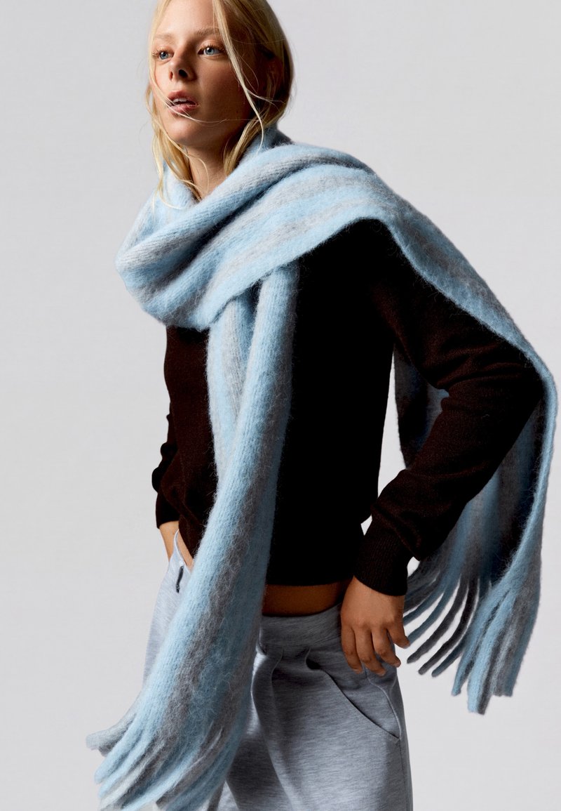 Femme blonde portant une grande écharpe pelucheuse bleu clair, un pull noir et un pantalon gris, debout les mains dans les poches.