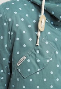 Morska zielona kurtka z wzorem w kropki w jasnoniebieskim kolorze, kapturem ze ściągaczem i kieszenią naszytą z przodu z metką.