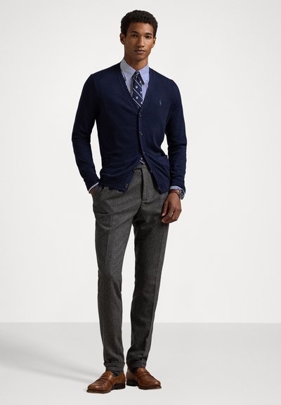 Navy wool cardigan over a striped shirt and tie, paired with tailored gray trousers and brown leather shoes. Classic, structured design.