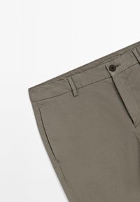 Grijze kaki broeksectie die de tailleband, riemlussen, knoopsluiting en voorzak toont op een witte achtergrond.