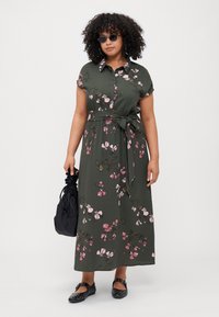 Green floral maxi dress with short sleeves, tie belt at the waist, and pink flower patterns. Paired with black sunglasses and a black bag.