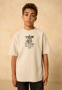 T-shirt beige a maniche corte con un motivo testurizzato, caratterizzata da un design grafico nero con la parola "TOKYO" e un'illustrazione di una tigre.