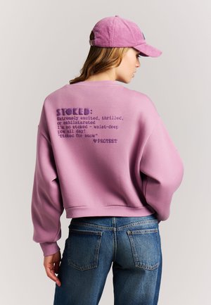 Woman wearing a pink sweatshirt with text on the back and a matching pink cap, paired with blue jeans, facing away from the camera.
