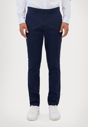 Mężczyzna ubrany w granatowe spodnie typu slim fit, białą koszulę i białe trampki stoi na jednolitym jasnym tle.