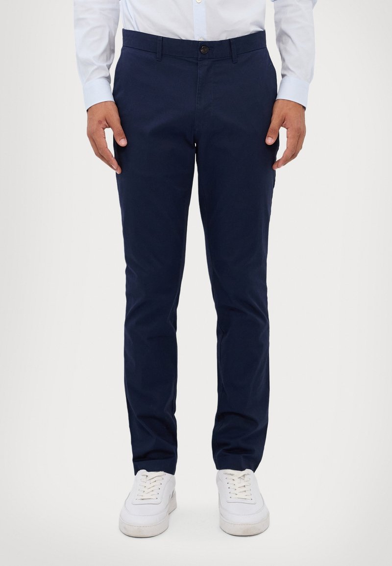 Homme portant un pantalon slim bleu marine, une chemise blanche et des baskets blanches, debout devant un fond clair uni.