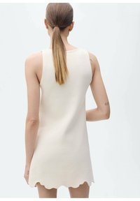 Abito mini senza maniche in tessuto a maglia beige, con orlo ondulato e texture liscia. Design minimalista con silhouette dritta.
