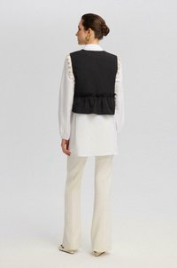 Chaleco negro con un dobladillo con volantes, llevado sobre una camisa blanca. El conjunto presenta pantalones crema acampanados y una textura suave. Los pendientes de oro añaden detalle.