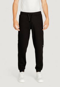 Black cotton sweatpants with an elastic waistband and drawstring, featuring side pockets and fitted cuffs. Paired with white sneakers.