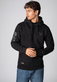 Zwarte hoodie van zacht materiaal, met een voorzak, een trekkoord capuchon en bedrukte tekst op de mouwen. Inclusief een logolabel aan de onderkant.