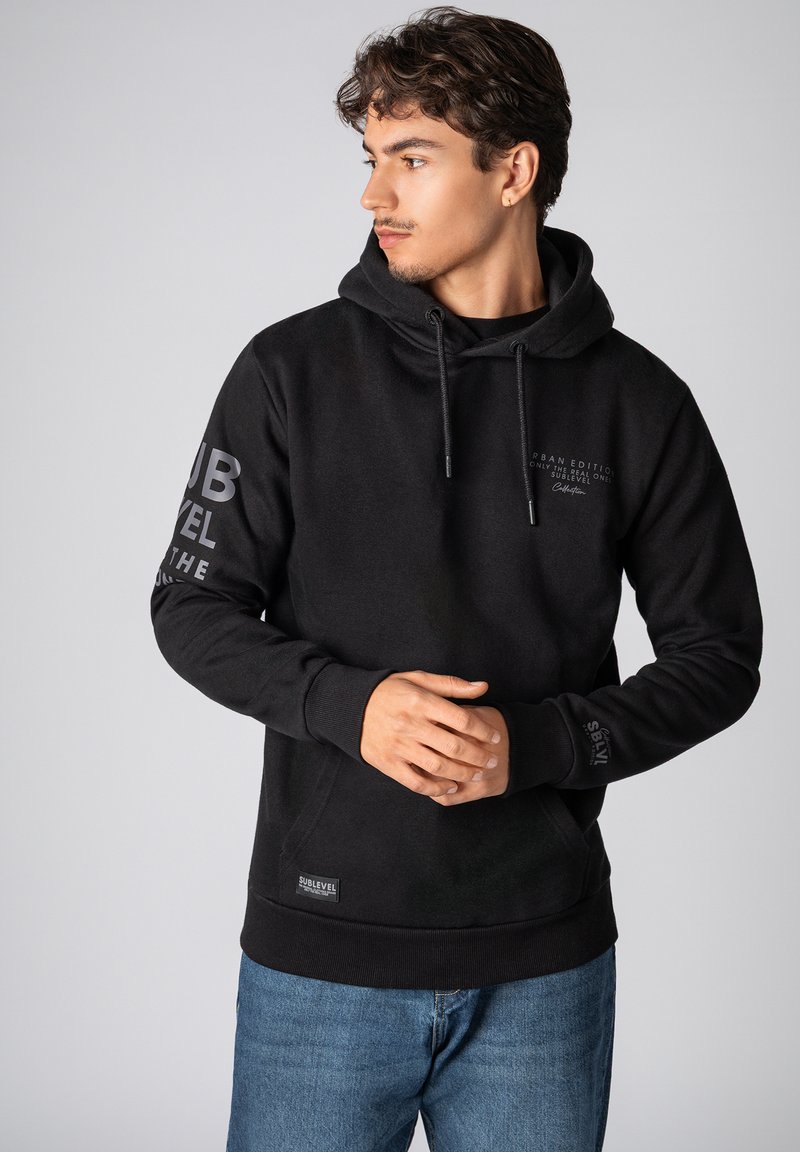 Zwarte hoodie van zacht materiaal, met een voorzak, een trekkoord capuchon en bedrukte tekst op de mouwen. Inclusief een logolabel aan de onderkant.