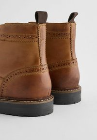 Botas de piel marrón hasta el tobillo con detalles de brogue, costuras decorativas y suelas de goma; cuentan con tiradores en la parte superior para facilitar su uso.