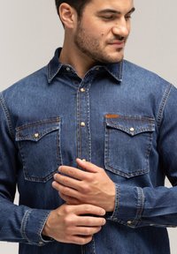Jeansowa koszula w ciemnym niebieskim kolorze, z dwiema kieszeniami na piersi, z zapięciem na guziki i kontrastowym przeszyciem. Posiada klasyczny kołnierzyk i długie rękawy.