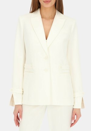 Kobieta nosząca dopasowaną białą marynarkę z wyciętymi klapami, dwoma guzikami, kieszeniami z klapkami i długimi rękawami, do białych spodni w komplecie.