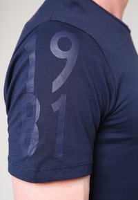 Námořnická modrá tričko s krátkým rukávem vyrobené z měkké bavlny, s velkým matným raženým grafickým motivem '1981' na horním levém rukávu.