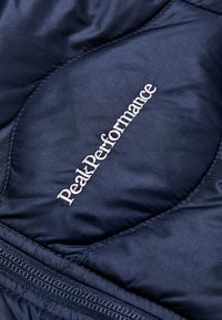 Giacca puffy blu navy con design trapuntato, texture liscia e logo bianco "Peak Performance" in contrasto sulla parte anteriore. Dettaglio della zip visibile.