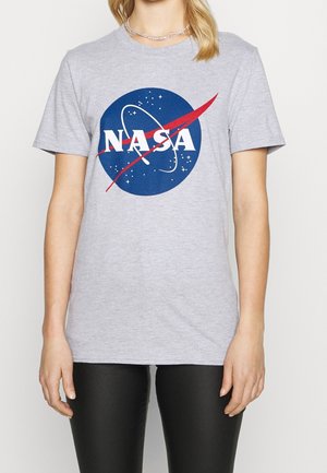 Kobieta nosząca jasnoszarą koszulkę z logo NASA w kolorach niebieskim, białym i czerwonym z przodu, do tego czarne spodnie.