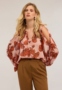 Blusa floreale di colore rosa chiaro con motivi di fiori rossi, con maniche a volant e scollo a V. Abbinata a pantaloni marroni a vita alta.