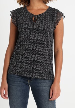 Mujer con blusa negra estampada de manga corta con mangas acampanadas y escote en forma de ojo de cerradura, combinada con vaqueros azules.