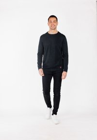 Camisa de manga larga verde oscuro con cuello redondo, combinada con jeans ajustados negros y zapatillas blancas. Presenta un sutil detalle del logo.