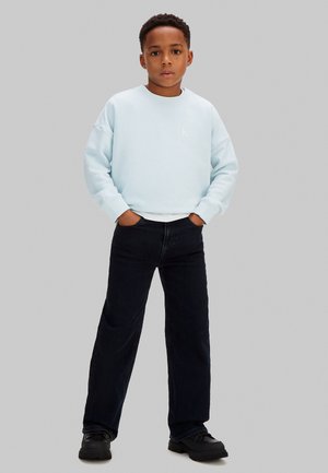 Ragazzo in piedi con le mani nelle tasche, indossa una felpa azzurra chiaro, pantaloni neri a gamba larga e scarpe nere su sfondo grigio.